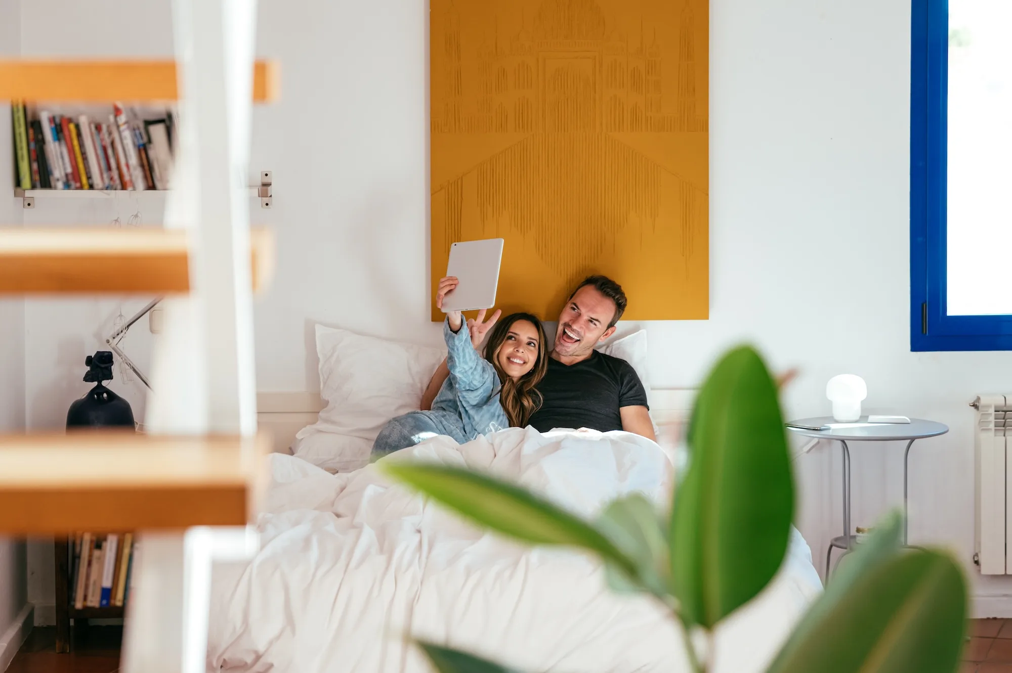 Cheerful man and woman smiling and gesturing V sign while taking selfie on comfortable bed in weekend morning